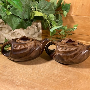 May include: Two brown ceramic teapots with lids, featuring a handle and spout. The teapots have a decorative design with a series of small, raised dots. The teapots are placed on a wooden surface, with green foliage in the background.