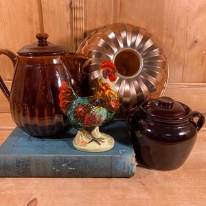 May include: A still life arrangement featuring a brown ceramic teapot, a copper bundt pan, a small brown lidded pot, and a colorful rooster figurine. The items are arranged on a blue book and a wooden surface.