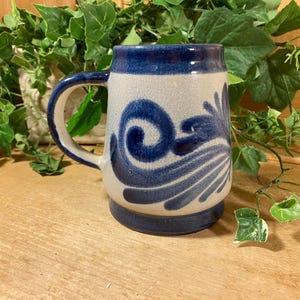 May include: A ceramic beer stein with a blue handle and base. The body is off-white with a blue floral design. The stein is cylindrical with a large handle and is sitting on a wooden surface.