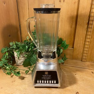 May include: Vintage Waring Pro Blender with a clear glass pitcher and black lid. The blender has a silver base with a black control panel and push buttons. The pitcher has vertical ridges. The blender is on a wooden surface.