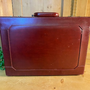 May include: A vintage burgundy leather briefcase with a metal clasp and a handle. The briefcase has a stitched design around the edges.