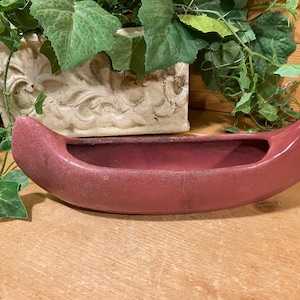 May include: A red ceramic canoe-shaped planter. The planter is empty and ready to be filled with plants.
