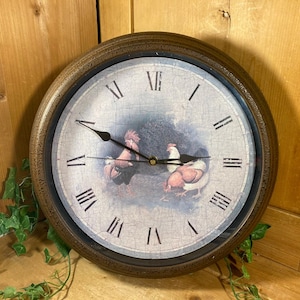 May include: Round wall clock with a distressed brown frame. The clock face features a rooster and hen illustration with Roman numerals. The clock hands are black.