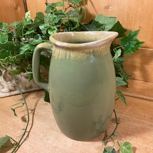 May include: A ceramic pitcher with a smooth, sage green glaze and a contrasting tan drip pattern around the rim. The vessel features a rounded body, a wide pouring spout, and a single curved handle attached to the side of the pitcher.