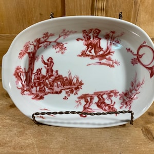 May include: Oval white serving dish with a red pastoral scene design. The dish has a scalloped edge and two handles. The design features figures, trees, and a hunting horn. The dish is displayed on a decorative metal stand.