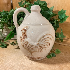 May include: Cream-colored ceramic jug with a handle and a rooster design. The jug has a rounded body and a narrow neck. The rooster is depicted in brown, with detailed feathers. The jug is likely vintage and adds a rustic touch to any kitchen or dining area.
