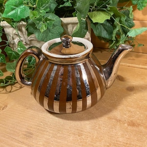 May include: A brown and silver striped teapot with a white lid. The teapot has a silver handle and spout.