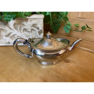 May include: A silver teapot with a lid and a handle. The teapot has a rounded body and a spout. The teapot is sitting on a wooden surface.