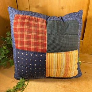 May include: Square patchwork pillow with a blue floral border. The pillow's front features four fabric squares: red plaid, navy blue with white dots, dark blue textured, and yellow and orange stripes. A cozy home decor item.