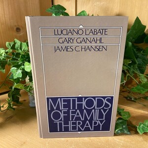 May include: A hardcover book titled "Methods of Family Therapy" with the authors' names: Luciano L'Abate, Gary Ganahl, and James C. Hansen. The book has a beige cover with a blue and white title. The background includes green foliage and a wooden surface.