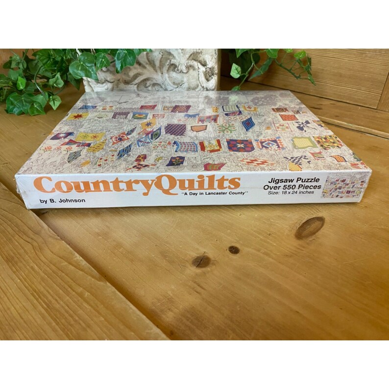 Może przedstawiać: Pudełko z puzzlem przedstawiające kolorowy obraz patchworkowych kołder z tytułem "Country Quilts" i tekstem "A Day in Lancaster County" autorstwa B. Johnsona. Na pudełku podano r&oacute;wnież, że puzzle zawiera ponad 550 element&oacute;w i ma wymiary 18 x 24 cali.