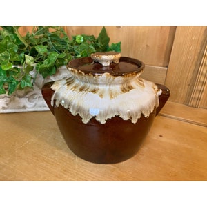 May include: A brown and white glazed ceramic crock pot with a lid. The crock pot has two handles and a drip glaze design.