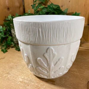 May include: Off-white ceramic planter with a textured bark-like band at the top and leaf pattern on the lower portion. The planter has a wide opening and is suitable for indoor or outdoor use. The neutral color complements various decor styles.