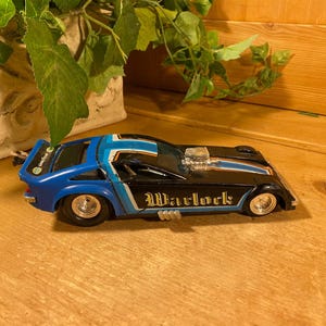 May include: A blue and black toy car with the word "Warlock" in gold lettering on the side. The car has a blue and white stripe design and chrome wheels. The car is sitting on a wooden surface.