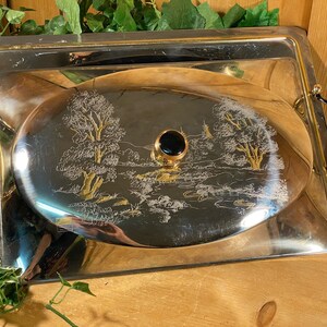May include: A silver serving tray with a gold rim and black handles. The tray has an oval-shaped lid with a black knob and an etched design of trees and a river.