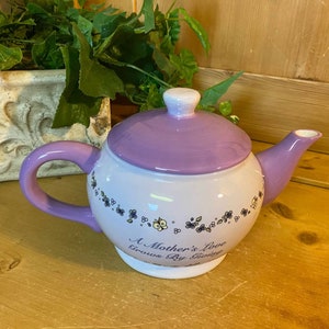May include: A purple and white teapot with a floral design and the text "A Mother's Love Grows By Giving".