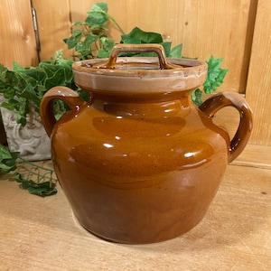 May include: A brown ceramic jar with a lid and two handles. The jar has a glossy finish and a light brown lid with a handle. The jar is sitting on a wooden surface.