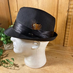 May include: A black fedora hat featuring a wide, smooth black satin ribbon band around the crown. A small, decorative cluster of brown and black patterned feathers is tucked into the side of the ribbon band, accented by a small metallic pin.