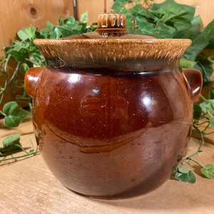 May include: A round, glazed ceramic bean pot with a dark brown body and a lighter, mottled tan rim. The pot features two side handles and a matching lid with a small, knob-like handle on top, showcasing a smooth, reflective finish throughout.