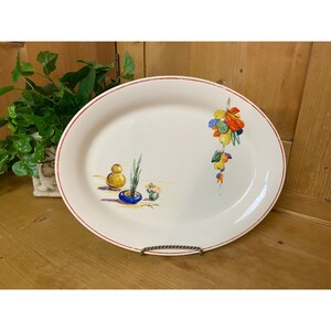 May include: A white oval serving platter with a red rim. The platter features a hand-painted design of a hanging bunch of colorful fruit and vegetables on the right side and a potted plant with a yellow gourd on the left side.