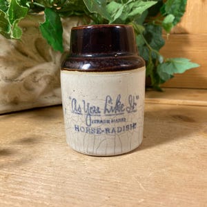 May include: A cylindrical stoneware crock with a dark brown glazed top and a cream-colored base featuring a crackled finish. The front displays blue printed text reading As You Like It, Trade Mark, and Horse-Radish in a decorative script font.