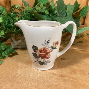 May include: Cream-colored ceramic pitcher with a gold rim and a curved handle. The pitcher features a floral design with red and yellow roses and green leaves. The pitcher is likely vintage or antique.