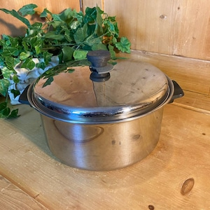 May include: A stainless steel pot with a lid and black handles. The pot is sitting on a wooden surface.