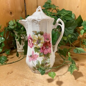 May include: A white porcelain coffee pot featuring a fluted body and a matching lid with a decorative finial. The exterior is adorned with a colorful floral pattern displaying pink, yellow, and red flowers with green leaves on the front surface.
