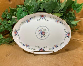 Può includere: Un vassoio ovale bianco con un bordo dorato e un motivo floreale. Il vassoio presenta un motivo floreale centrale con fiori blu e rosa e foglie verdi. Il bordo del vassoio è decorato con un motivo ripetuto di fiori blu e rosa e foglie verdi.