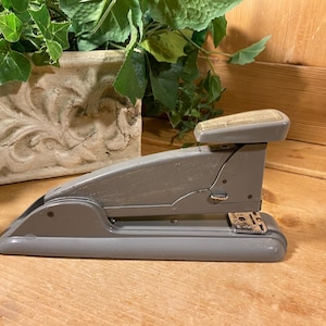 May include: A gray metal stapler with a silver top and a silver staple remover. The stapler is on a wooden surface.