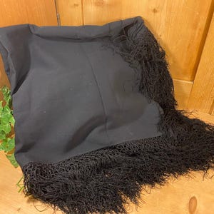 May include: A black shawl with a fringed edge. The shawl is folded and appears to be made of a soft fabric. The fringe is long and adds texture to the garment. The shawl is displayed on a wooden surface.