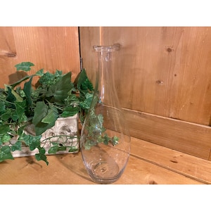 May include: A clear glass vase with a rounded bottom and a narrow neck. The vase is empty and sitting on a wooden surface.