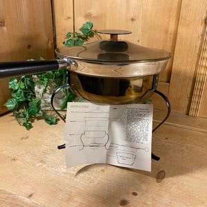 May include: A vintage fondue pot with a shiny metal finish, black handle, and lid. The pot sits on a black metal stand with a paper instruction sheet in front. The background is wooden.