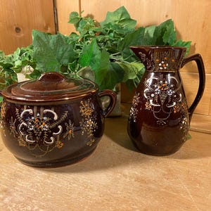 May include: A dark brown ceramic pitcher and covered pot set. Both pieces are decorated with white, orange, and gold floral patterns. The pitcher has a handle and spout, while the pot has two handles and a lid.