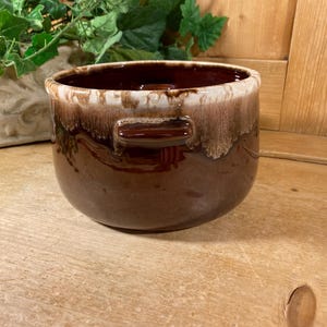 May include: A round ceramic bowl with two small side handles, featuring a deep glossy brown glaze on the lower portion and a contrasting creamy white drip glaze around the upper rim, creating a distinct layered color effect on the exterior.