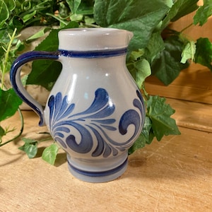 May include: A ceramic pitcher with a white and blue floral design. The pitcher has a blue handle and rim. The body of the pitcher is a light gray color. The floral design is a darker shade of blue. The pitcher is approximately 6 inches tall.