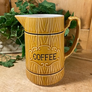 May include: A tan ceramic coffee pitcher with a wood grain design. The pitcher has a handle and a spout. The word "COFFEE" is displayed in a decorative font on the front of the pitcher. The pitcher is sitting on a wooden surface.