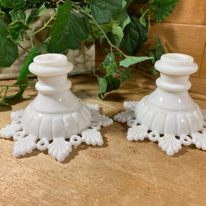 May include: Two white milk glass candle holders. Each has a round base with a decorative, openwork design and a fluted body. The top has a circular opening for a candle. The candle holders are on a wooden surface.