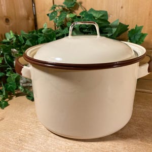 May include: Cream-colored enamel pot with a matching lid and brown trim. The pot has two side handles and a handle on the lid. The pot is sitting on a wooden surface with green foliage in the background. A vintage kitchenware item.