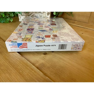 Może przedstawiać: Pudełko na puzzle z obrazkiem kolorowych patchworkowych kołder. Pudełko jest białe z czerwonymi, białymi i niebieskimi akcentami. Tekst na pudełku brzmi "Great American Puzzle Factory" i "Jigsaw Puzzle #974".
