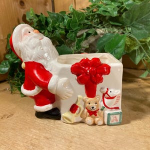 May include: A ceramic Christmas planter featuring Santa Claus in a red suit with white trim. The planter is white with a red bow and toy illustrations. A teddy bear, rocking horse, and bell are visible.