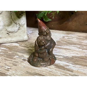 May include: A bronze figurine of a gnome hugging a dog. The gnome is wearing a red hat and the dog is brown. The base of the figurine says "Best Friends".