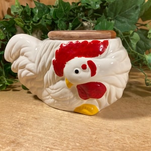 May include: A set of three ceramic mugs with a speckled, cream-colored glaze. Each mug features a different hand-painted design: a blue and green leaf, a yellow flower, and a red berry. The mugs have a simple, rounded shape.
