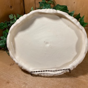 May include: A white, oval-shaped ceramic serving dish with a scalloped edge. The dish is supported by a dark metal stand and is surrounded by green foliage. The dish is empty and sits on a wooden surface.
