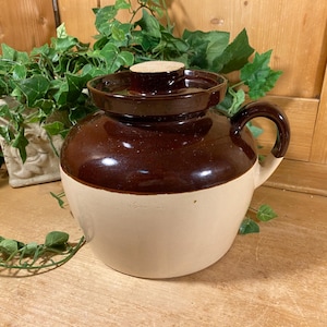 May include: A two-toned ceramic crock featuring a glossy dark brown glazed upper section and a smooth cream-colored lower base. The vessel includes a matching dark brown lid with a central knob and a single curved handle on the side for gripping.