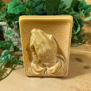 May include: A ceramic wall plaque with a raised design of two hands clasped in prayer. The plaque is a light brown color with a textured surface.