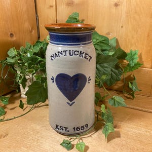 May include: A cylindrical stoneware crock with a light gray speckled finish, featuring a dark blue heart graphic in the center. The text NANTUCKET is printed above the heart, and EST. 1659 is printed below. The rim is accented with a dark blue band.