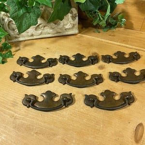 May include: Eight antique brass drawer pulls with a decorative, ornate design. The pulls have a curved shape with a raised center and are in good condition.