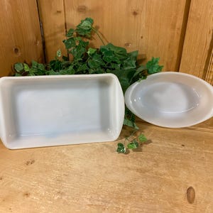 May include: Two white ceramic baking dishes. One is rectangular with rounded corners and handles, and the other is oval with handles. Both dishes are empty and sit on a wooden surface with green foliage in the background.