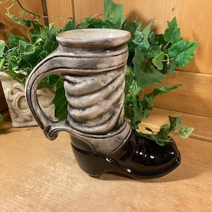 May include: A ceramic boot-shaped planter with a black glossy toe and a gray and white upper section. The boot has a large handle and is filled with greenery. The planter is on a wooden surface.